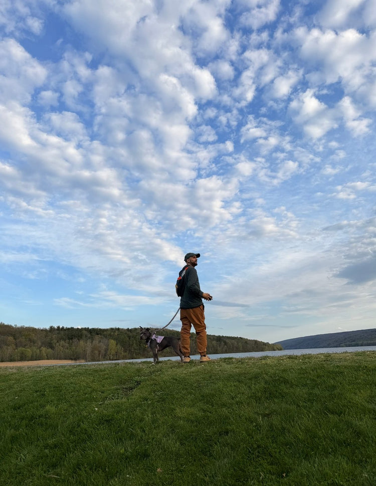 Tillie standing on a grassy lakeshore on her bungee leash beside a man in a cap, under a wide cloudy sky