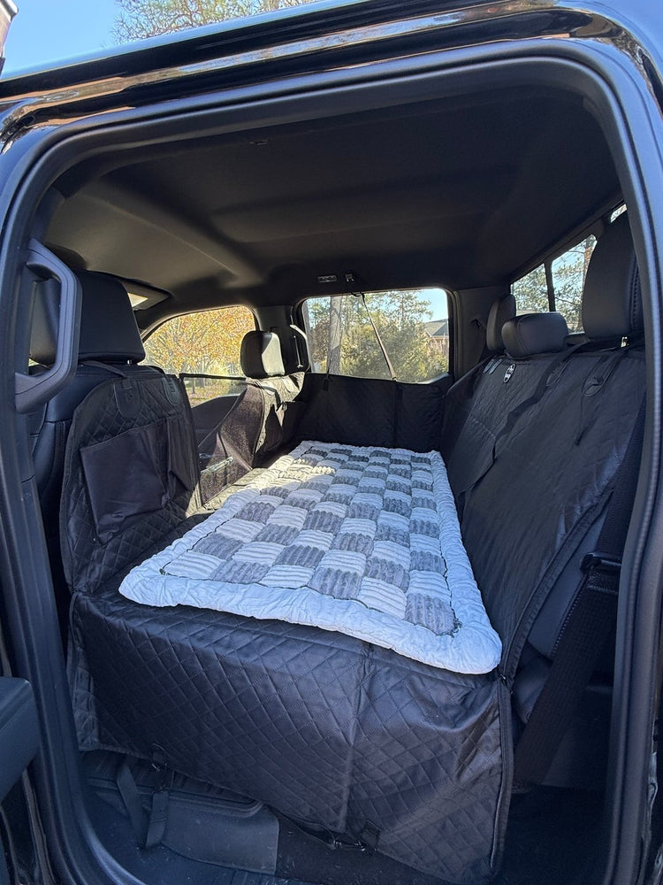 A plush gray and white Team K9 backseat bed lying on a black Team K9 hard bottom seat cover in the back of a truck